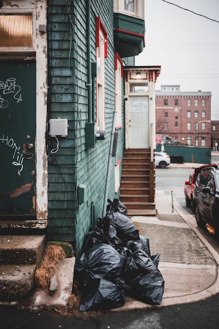 A narrow urban alleyway beside a green wooden building with weathered, uneven shingles. Black plastic rubbish bags are piled at the base of a short flight of wooden stairs leading to a white door with a small window above, supported by a white and red striped awning. The bags appear filled with discarded waste, and some are stacked against the building's exterior wall, nearby a patch of exposed brown ground and a concrete step. To the left, part of a garage door or shutter with graffiti and peeling paint is visible. In the background, parked cars line the street, with a large brick building further away under an overcast sky. The scene captures a typical on-site waste disposal area, which could be handled by private rubbish removal services such as those offered by Rubbish Removal Twickenham, reflecting an alternative method of local waste management outside council collection. The environment suggests a typical residential or commercial property’s alley used for rubbish storage before collection or disposal.