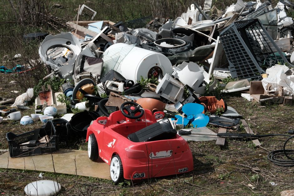 A large pile of mixed household waste and discarded items scattered across an outdoor natural environment, including a small red plastic toy car with black side panels and white wheels positioned in the foreground. Surrounding the toy are various objects such as broken appliances, plastic containers, utensils, and electronic parts with visible textures of hard plastic, metal, and ceramic. The waste features a variety of colors, predominantly white, black, grey, and brown, with some items exhibiting worn or damaged surfaces. The background consists of sparse vegetation and overgrown grass, indicating an outdoor setting that appears to be an informal dumping site. Some larger items, like a white washing machine drum, stands out among the clutter, and an assortment of smaller items such as bowls and baskets are intermingled within the pile. The scene is illuminated by natural daylight, highlighting the textures and the disorderly nature of the waste, which could be associated with private rubbish disposal or alternative waste handling services like those offered by Rubbish Removal Twickenham.