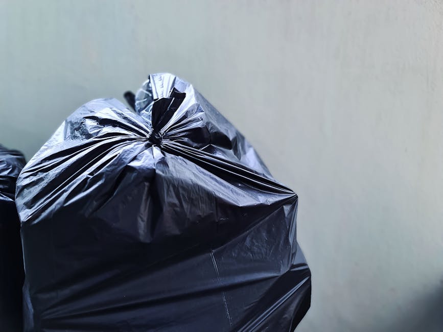 Close-up image of several large black plastic rubbish bags tightly sealed with knots, showing crinkled and reflective surfaces. The bags are placed on a surface likely within an indoor or sheltered environment, with a plain, light-colored wall in the background. The bags contain waste materials, possibly household or general rubbish, prepared for collection or disposal. The lighting is even, highlighting the glossy texture of the plastic. This scene exemplifies typical private waste handling or on-site rubbish collection, aligning with services offered by Rubbish Removal Twickenham for efficient and independent rubbish removal. The focus on sealed bags and neutral background creates a professional and factual impression suitable for a rubbish removal service context.