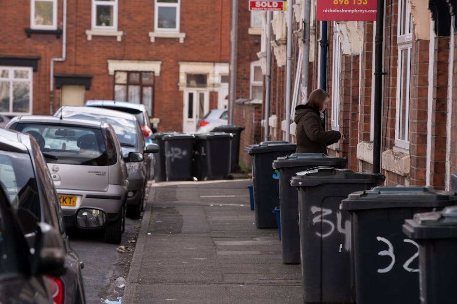 A row of black wheelie bins lined up along a residential pavement adjacent to a brick building with multiple white-framed windows. The bins are positioned close to the wall, each with a closed lid, some displaying white spray-painted numbers on their sides. A woman dressed in a brown coat is seen placing waste into one of the bins, standing on the sidewalk next to the building. Behind her, additional bins are visible further along the street, which is lined with parked cars of various sizes and colors, including silver, dark grey, and light blue. The street appears to be in a terraced housing area with red-brick facades and small front yards or steps leading to entrance doors. The scene is illuminated by natural daylight, and the overall setting suggests a typical urban residential environment where private rubbish collection services, like those offered by Rubbish Removal Twickenham, may be involved in waste management or on-site clearance activities.
