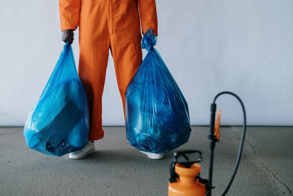 A person wearing an orange work jumpsuit and gloves is standing on a plain indoor surface, holding two large, semi-transparent blue plastic rubbish bags filled with waste. The bags are slightly creased and bulging, indicating they are tightly packed with debris. The individual is positioned against a neutral, light-colored background, with only midsection and legs visible. Nearby, a small orange waste sprayer with a black hose and handle is placed on the ground, suggesting an environment related to waste handling or cleaning. This scene exemplifies on-site waste collection or disposal activities, aligning with private rubbish removal services such as those offered by Rubbish Removal Twickenham, particularly in contexts demanding independent clearance of household or commercial waste.